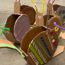 Load image into Gallery viewer, A group of handmade cardboard snail crafts arranged on a concrete floor. Each snail is made from brown corrugated cardboard with a spiral shell decorated with colorful patterns like stripes, polka dots, and floral prints.